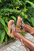 A person wearing carved wooden platform sandals with rhinestone-embellished straps, photographed outdoors against a background of green tropical plants and a paved walkway.