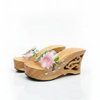 A pair of wooden wedge mule sandals with carved details and a clear strap decorated with a pink and green flower, set against a plain white background.