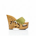 A single wooden wedge sandal with a carved floral design on the heel, a light brown wood platform, and a textured olive green strap adorned with three gold-tone rivets, shown against a plain white background.