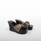 A pair of women's wedge mule sandals with glossy black cutout platforms and straps featuring a leopard print pattern in brown, beige, and black.