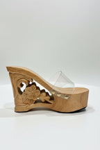 A single wooden wedge sandal with floral carvings on the heel, a clear plastic toe strap, and small gold-tone rivets, displayed on a white table against a brown curtain background.